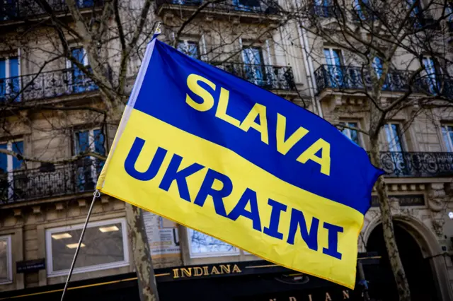 Quốc kỳ Ukraine cùng khẩu hiệu Slava Ukraini (Vinh quang thuộc về Ukraine) tại một cuộc biểu tình do các tổ chức ủng hộ Ukraine kêu gọi, vào dịp trước ba năm kể từ khi Nga xâm lược Ukraine, trên Đại lộ du Temple (giữa Place de la République và Place de la Bastille) ở Paris, Pháp, ngày 23/2/2025.