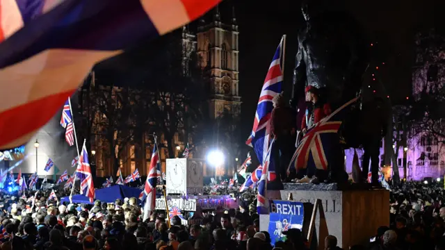 Tłum ludzi zgromadzonych nocą na placu Parlamentu w Londynie macha flagami Union Jack.