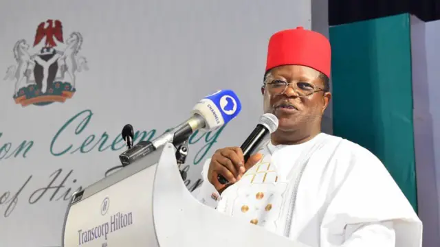 David Umahi tsaye yana jabawani sanye da fararen tufafi da gariya mai aiki a gabanta da jar dara a gaban abin d ake jawabi a kansa na hotel ɗin transcorp yana rike da abin magana baƙi mai kan ruwan toka da kuma abin maganar gidan talbijin na channels.