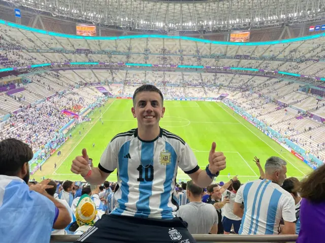 Fotoğrafta Arjantin forması giyen bir adam görülüyor. Mutlu görünüyor, kameraya gülümsüyor ve başparmağını yukarı kaldırıyor. Arka planda bir futbol stadyumu var. Etrafında diğer Arjantin taraftarları bulunuyor.
