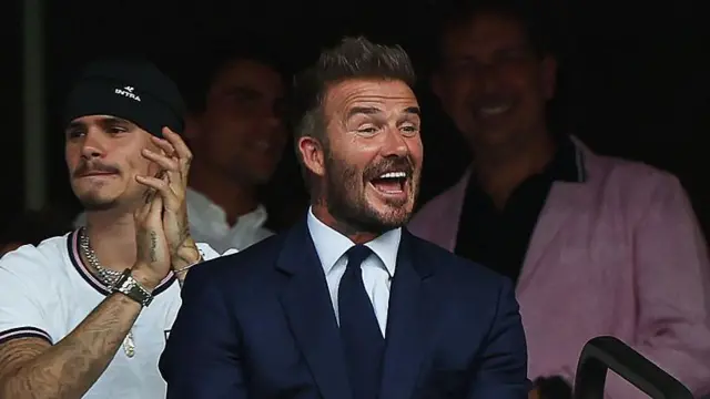 O ex-jogador de futebol inglês David Beckham sorri durante a partida das quartas de final da Copa do Mundo de 2026 entre Noruega e Inglaterra, no Estádio de Miami, em Miami Gardens, no dia 11 de julho. Ele veste um terno azul, camisa branca e gravata azul.