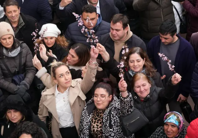 Protestoda çok sayıda kadın ellerinde çiçek tutuyor, hava soğuk ve mont giymişler.