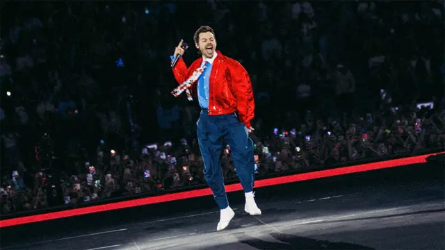 Harry Styles ma na sobie granatowe spodnie, jasnoniebieską koszulę z białym kołnierzykiem i krawatem w kwiatowy wzór oraz czerwoną kurtkę. Skacze na scenie, a za nim widać fanów obserwujących występ.