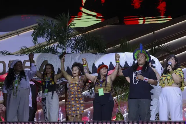 Jovens, alguns deles com itens indígenas, seguram e erguem as mãos em palco