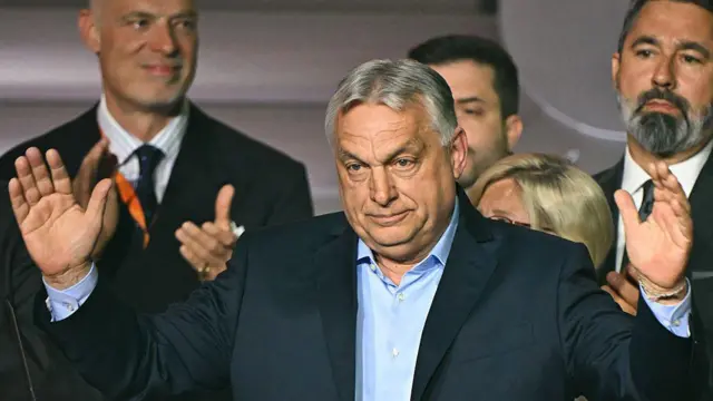 Orbán Viktor, Magyarország volt miniszterelnöke két oldalon feltett kézzel. Mögötte három másik ember részlegesen látható.