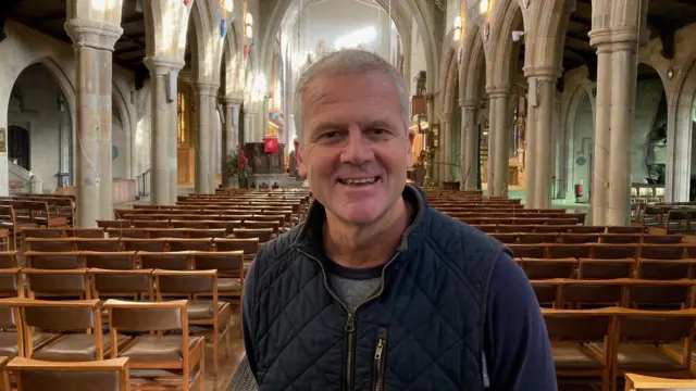 Un hombre vestido con una chaqueta acolchada azul dentro de una catedral con filas de asientos de madera al fondo.