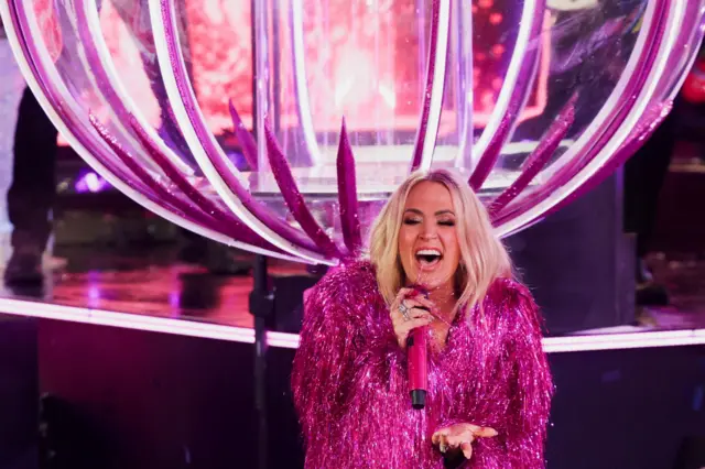 Carrie Underwood, mulher loira com roupa rosa, cantando em palco