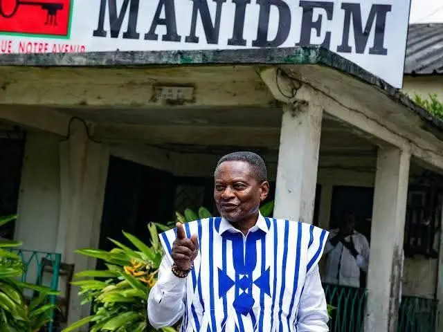 Georges Anicet Ekane, porte une chemise blanche avec un par-dessus blanc bleu, lève le doigt de sa main droit. Derrière lui une pancarte sur laquelle est écrit "MANIDEM" est posée sur un bâtiment.