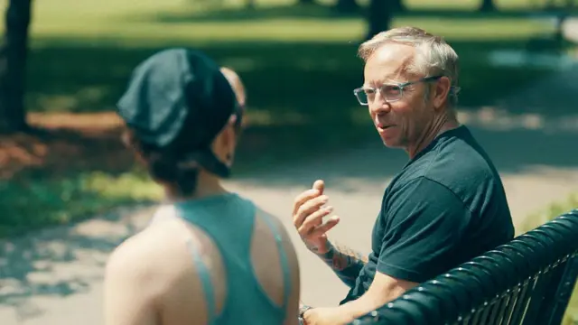 পার্কের বেঞ্চে বসে স্কয়ার এবং লুসি কথা বলছে। স্কয়ার গাঢ় রঙের টি-শার্ট পরে আছে এবং লুসি ক্যাপ এবং নীল রেসার-ব্যাক টপ পরে আছে।