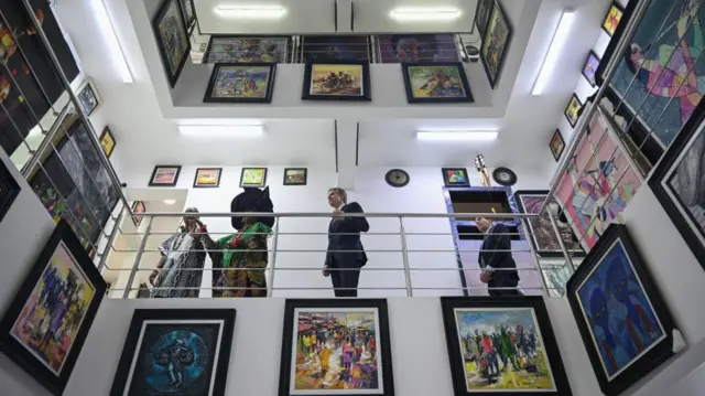 Photographed from below, Mama Nike and Antony Blinken stand on an upper-level walkway inside a brightly lit gallery, alongside a number of other people. They are surrounded by framed artworks displayed on the walls and balcony railings. In the foreground and background, colourful paintings of figures and abstract scenes cover both floors of the white interior, all the way up to the high ceiling.