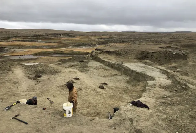 A wide view shows the barren brown landscape of the Badlands in South Dakota, with two people in the foreground on a dig with tools around them, on a cloudy grey day.