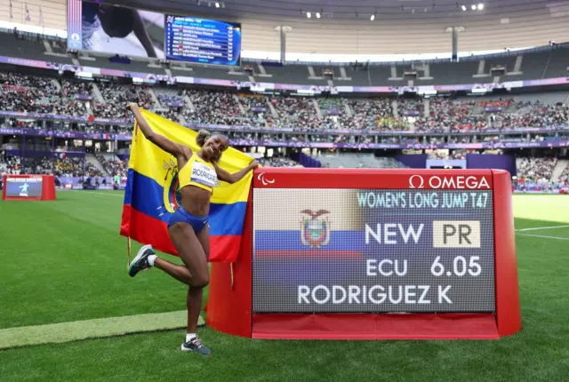 La atleta Kiara Rodríguez, de Ecuador, festeja junto a una pantalla que muestra su récord paralímpico en salto largo.