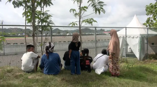 Tujuh orang warga menonton MotoGP dari pekarangan Sibawaih. Lima di antaranya duduk di rumput, sementara dua lainnya menonton sambil berdiri. Meski terhalang oleh pagar, mereka tetap bisa menyaksikan motor yang melintas dengan jelas.
