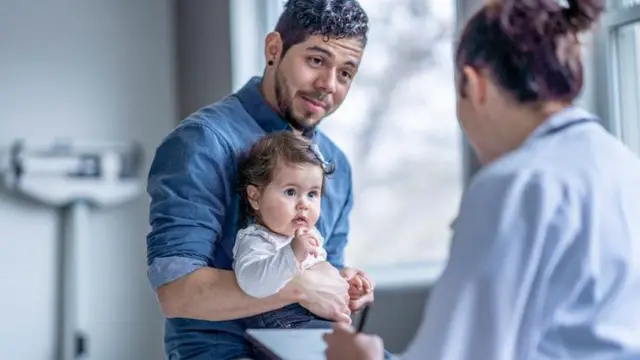 Homem com bebê no colo, observado por uma médicaapostar na betwayconsultório