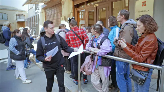 Un hombre con una sudadera negra con capucha y vaqueros está repartiendo documentos a las personas que hacen cola frente a un edificio oficial cerca del Kremlin.