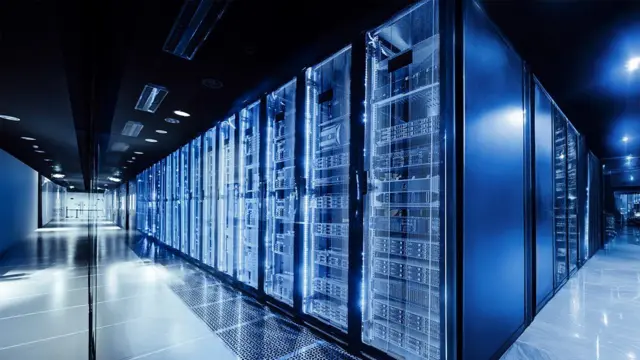 Imagen de un centro de datos. Un pasillo se bifurca hacia la izquierda y hacia la derecha. En el medio se ve una larga hilera de torres de color azules dentro de las cuales parece haber máquinas de procesamiento de datos.