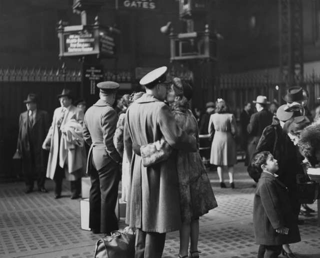 Una pareja que se besa y está abrazada se encuentra en el centro de la foto. Él lleva un gorro y un abrigo largo, ella un tocado negro en la cabeza y un abrigo elegante. Es una estación de trenes en la que se ven otras personas. Hay otro uniformado de espaldas