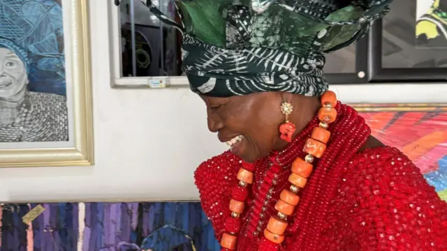 Mama Nike is shown in profile, laughing as she looks down. She is wearing the same bright red beaded garment, large green patterned headwrap and matching red and orange bead jewellery. Behind her are framed artworks displayed on white gallery walls, including a black-and-white portrait on the left and a colourful painting partially visible in the background.
