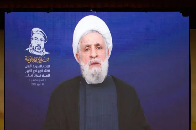 نعیم قاسم در حال سخنرانی، روی یک صفحه نمایش، با لباس سیاه و عمامه سفید
