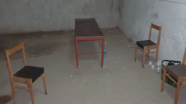 Фото помещения с голыми бетонными стенами и полом, тремя стульями, столом и бутылками на полу 
