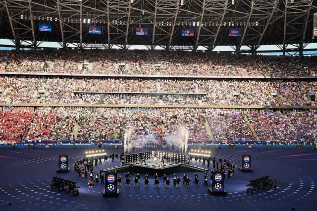 Picture of the stage at the Puskas Arena, Budapest. A large stand of crowds is seen in the backgorund of a stage in the middle of the pitch.