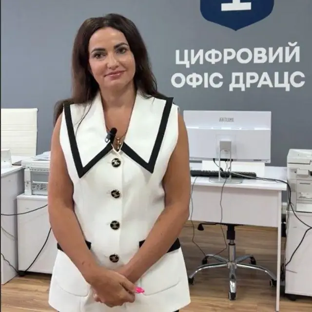 жінка в біло-чорному жакеті з чорним волоссям стоїть і дивиться в камеру 