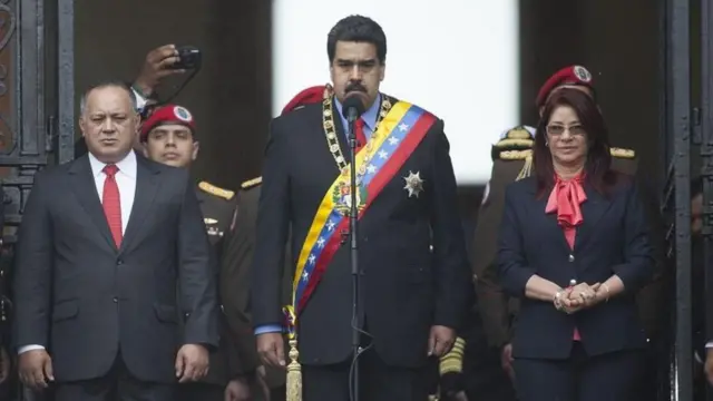 Flores stands next to Maduro and another man, all in very formal attire and with stern expressions, with men in military uniform behind them.