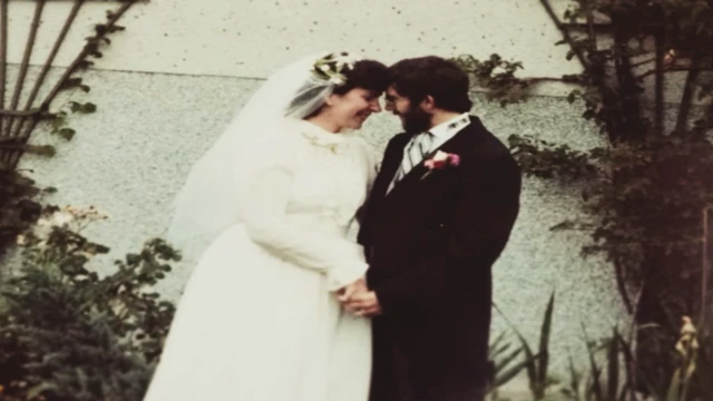 Lee McKenzie is a white woman with dark hair and is wearing a white long-sleeved wedding dress and a veil. Kenner Jones is wearing a black morning suit with a black, grey and white striped tie and a pink flower on the lapel. They are standing in front of a white wall with plants to either side. They are holding hands and smiling at each other.