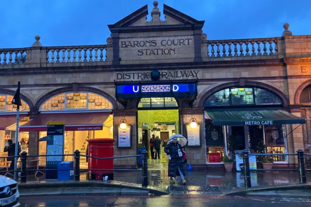 El exterior de la estación Barons Court muestra un cartel del metro y una mujer con paraguas.