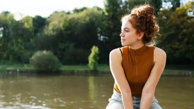 امرأة ترتدي قميصاً بنياً بدون أكمام وبنطال جينز أزرق فاتح، شعرها أحمر مربوط إلى الأعلى، تجلس على حافة تظهر خلفها بحيرة.