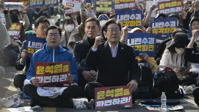 首爾街頭身穿黑色外套的韓國民主黨黨魁李在明（中）拿著反對尹錫悅標語牌帶領支持者叫喊口號（22/3/2025）