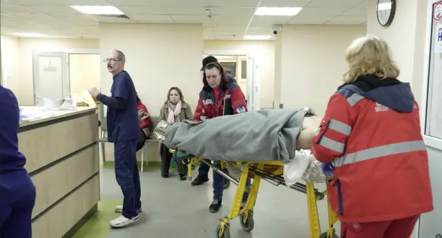 На фото - приймальне відділення лікарні Мечникова. В центрі - ліжко-каталка з пацієнтом, накритим ковдрою та двомами медиками-жінками швидкої, які його транспортують. 