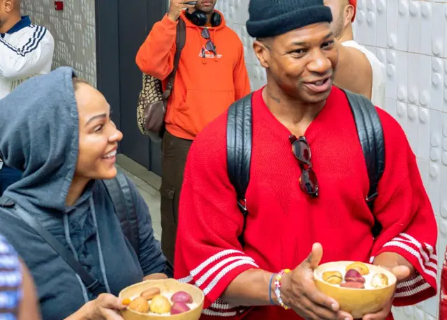 Les acteurs américains Meagan Good et Jonathan Majors sont accueillis à l'aéroport international Conakry Gbessia. On leur a offert des noix de kola. Les noix de kola ont une grande importance culturelle en Afrique de l'Ouest, où elles symbolisent l'hospitalité, la paix, l'amitié et la vie. Elles sont utilisées dans des rituels sociaux importants tels que les mariages, les funérailles et les négociations. Le couple sourit, discute et porte des vêtements décontractés