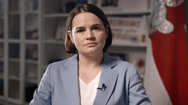 Светлана Тихановская в своем офисе в Вильнюсе во время интервью для Би-би-си в 2025 году. Она в серо-голубом пиджаке на фоне бело-красно-белого флага и стеллажа белого цвета