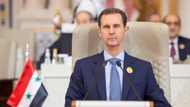 Basar al-Assad está sentado ligeiramente inclinado em sua cadeira, com uma expressão séria no rosto e a bandeira da Síria visível no chão próximo. Ele está vestindo um terno azul e gravata azul.  