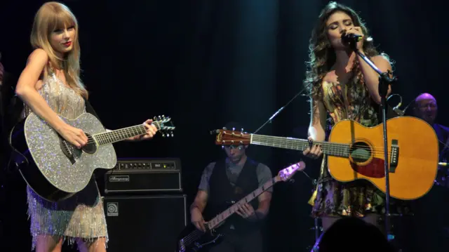A imagem mostra duas cantoras se apresentando juntas em um palco, cada uma com um violão acústico. À esquerda, Taylor Swift veste um figurino brilhante com franjas prateadas e olha em direção ao público, enquanto Paula Fernandes, à direita, canta ao microfone usando um vestido estampado.