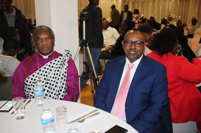 Fidel Twagira (iburyo),mw'ikoti y'ubururu yicaye ku meza iriko ibikoresho vyo kurya. I buryo bwiwe hicaye umugabo yambaye umwambaro uranga abapasitori canke abapatiri yarengejeko umutama w'akaranga. 