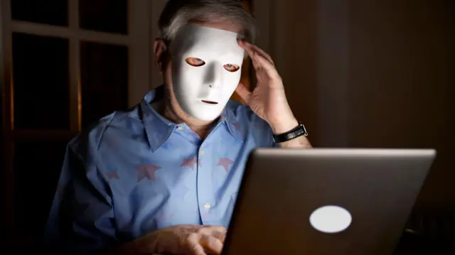 شخص يرتدي قناع ويجلس أمام شاشة لابتوب