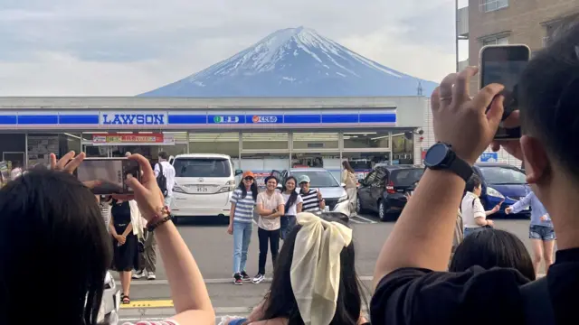 日本山梨县富士河口湖町一家便利店外游客以便利店与富士山为背景拍照留念（28/4/2024）
