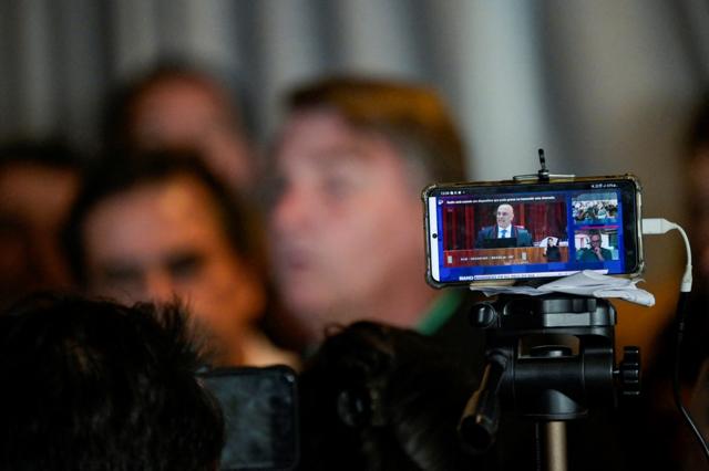 No primeiro plano, celular com imagemtigre 777 linkAlexandretigre 777 linkMoraes; no fundo, Bolsonaro dá entrevista