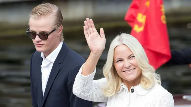 Kobieta w białej sukni i blond włosach uśmiecha się, a obok niej stoi młody chłopiec