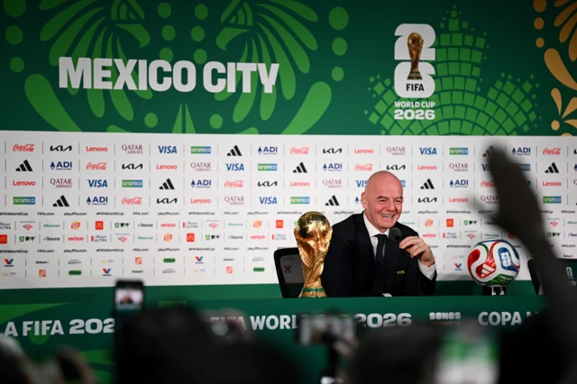 Infantino está sentado y sonríe. Sobre la mesa, al frente de él, está la copa del Mundial y un balón de fútbol. Detrás de él hay una pared con marcas de patrocinadores y en letras grandes dice: Mexico City. FIFA World Cup 2026. Se ve la mano de un periodista levantada