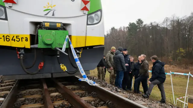 Biało-żółto-zielony pociąg stoi na torach. Do pociągu przymocowana jest taśma policyjna. W tle widać grupę rozmawiających mężczyzn.