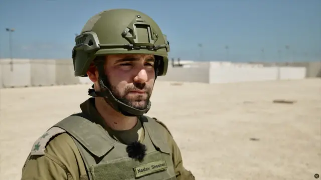 Nadav Shoshani is wearing a khaki military uniform, including a helmet. His name is written on the protective vest across his chest. He appears to be in his 20s or 30s and has dark eyebrows and a beard.
