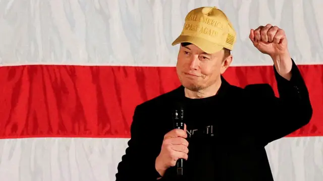 Elon Musk vestindo um blazer preto e um boné amarelo onde se lê "Make America Great Again", slogan da campanhajogo roleta da sorteTrump, com a mãojogo roleta da sortepunho e segurando um microfone na outra mão. 