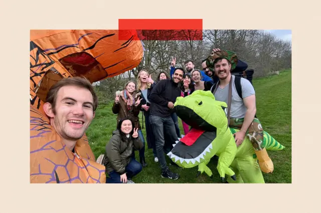 A group of people stand in a park, some of them wearing dinosaur costumes.