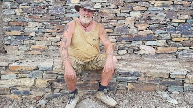 Darren "Badger" Edge lleva camiseta, pantalones cortos y botas de trabajo. Sonríe y está sentado en un muro de piedra.