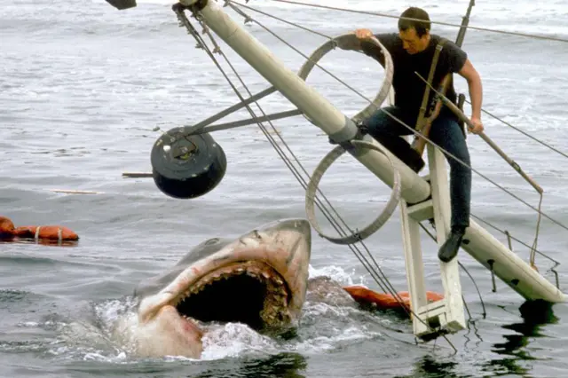 No set de gravação de 'Tubarão', o ator Roy Scheider (1932-2008) aparece sobre os restos de um barco que afunda, enquanto o tubarão o espera com a boca aberta na água