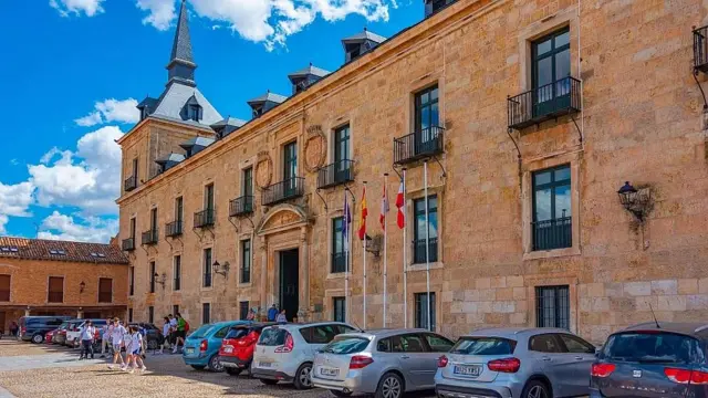 Fachada del Parador de Lerma . Es una construcción antigua de piedra color terracota de tres pisos con balcones de hierro en el piso más alto. Al frente hay varios autos aparcados y se ve a huéspedes del parador saliendo a la calle