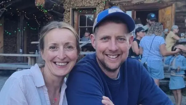 Caroline veste uma camisa clara e Gareth um moletom azul escuro. Ambos estão sorrindo. Ao fundo, é possível ver um pequeno grupo de pessoas, incluindo uma criança